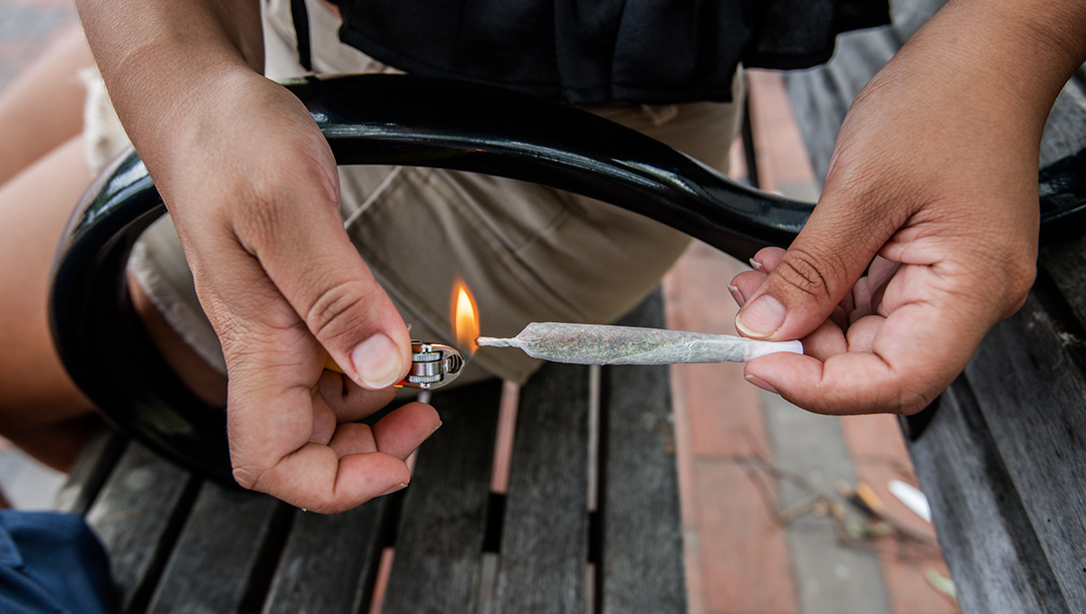 よりリベラルな大麻法がスパイス／K2の過剰摂取を減少させる: 公園のベンチでカンナビスジョイントに火をつける女性の手元