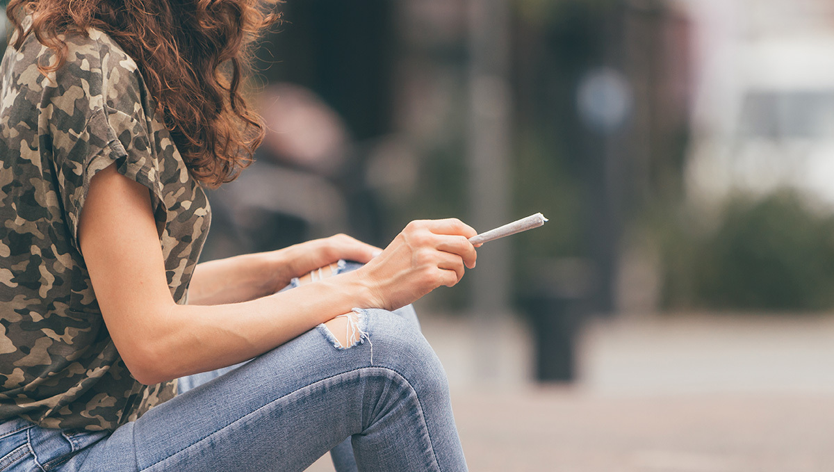 Spain to Legalize Medical Plants Sales This Year: A young woman in denim an khaki shirt sitting on a curb with an unlit joint in hand
