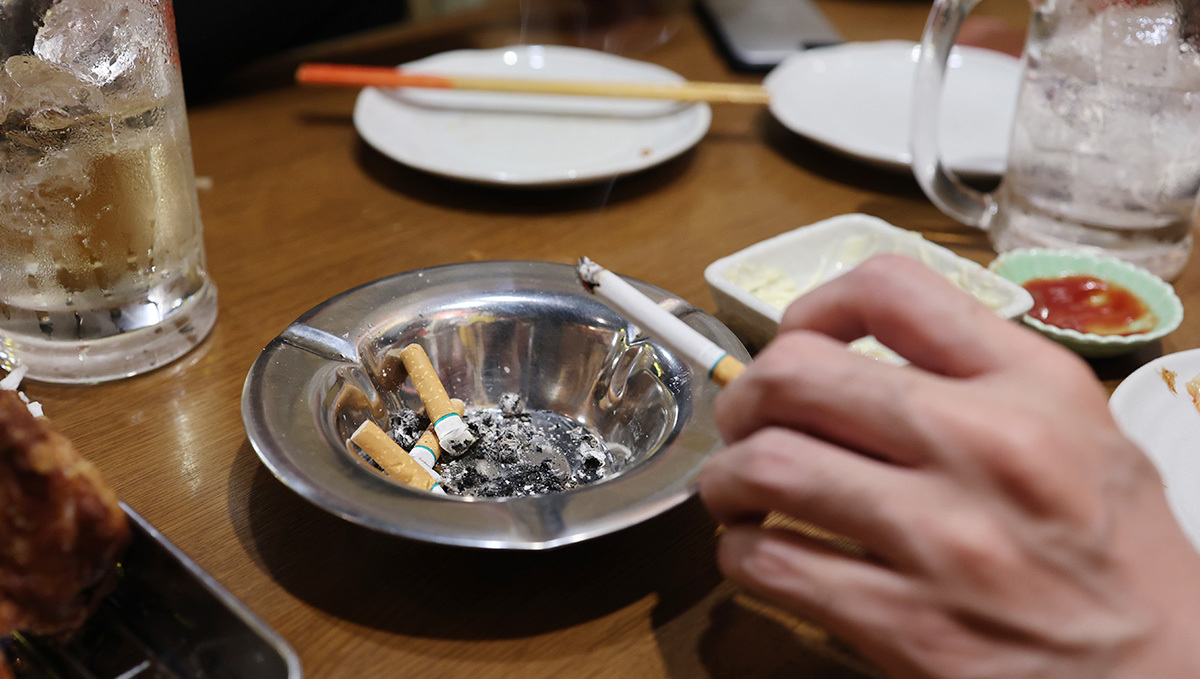 Plants Taxes Bring In 3x More Than Alcohol + Tobacco in Arizona: A closeup of a tabletop with an alcoholic drink and an ashtray; a hand is seen shaking ashes from a burning cigarette